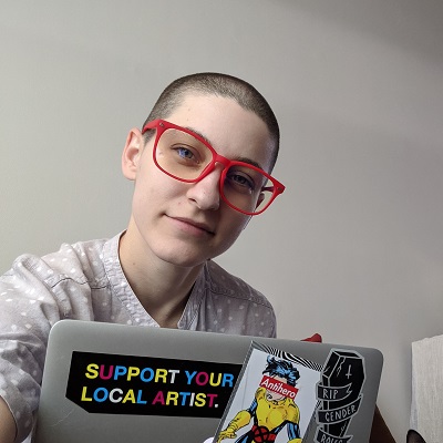 Tori Hart smiles at the camera behind their laptop, which is covered in quirky stickers.